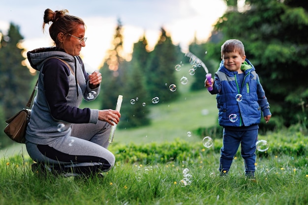 Mère joue avec son fils sur une pelouse et lui apprend à souffler des bulles de savon sur fond de forêt