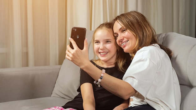 Une mère joyeuse et sa fille adolescente en t-shirts noir et blanc font un selfie souriant et assis sur un canapé dans le salon à la maison