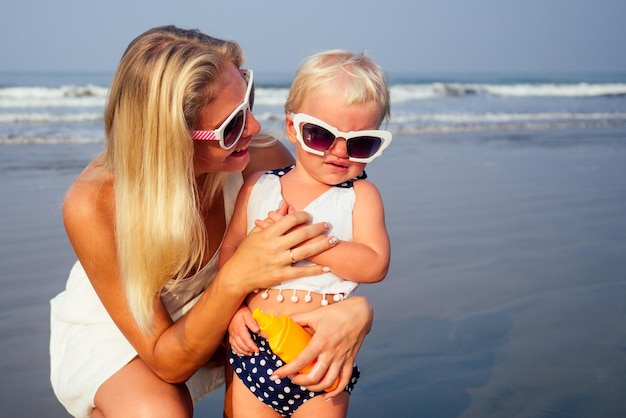 Mère en lunettes élégantes et robe blanche met un flacon pulvérisateur orange SPF sur bébé nouveau-né fille d'un an un écran solaire sur la plage