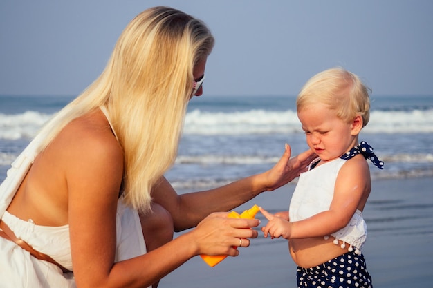 Mère en lunettes élégantes et robe blanche met un flacon pulvérisateur orange SPF sur bébé nouveau-né fille d'un an un écran solaire sur la plage
