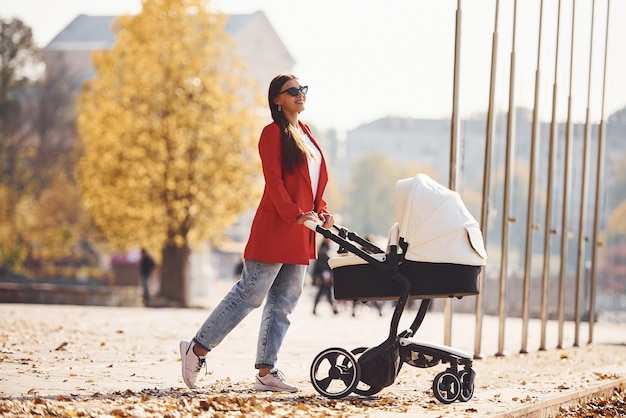 Une mère en manteau rouge se promène avec son enfant dans le landau du parc à l'automne.