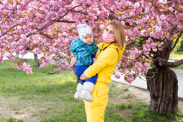 Mère en masque médical et petit garçon fils s'amusant dans le parc du printemps près de l'arbre en fleurs de sakura rose concept de printemps