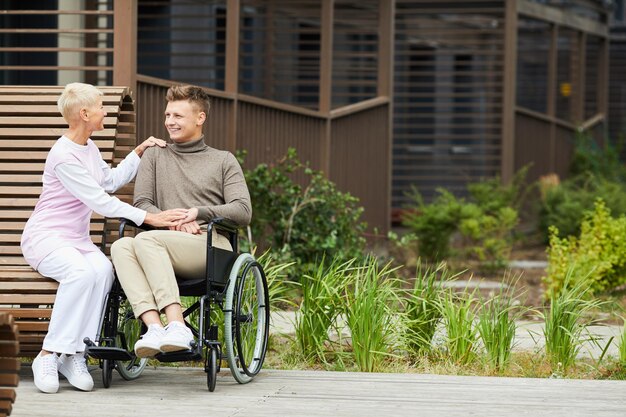 Mère mature souriante assise sur un banc et caressant son fils en fauteuil roulant pendant qu'elle lui rendait visite en reco