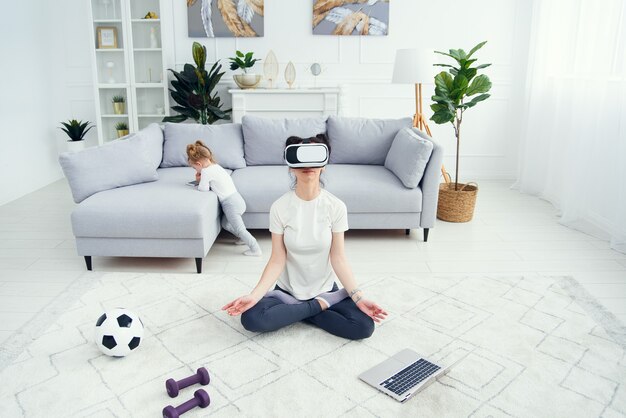 Mère méditant en position de yoga lotus utilise des lunettes vr pendant que sa fille regarde des dessins animés sur