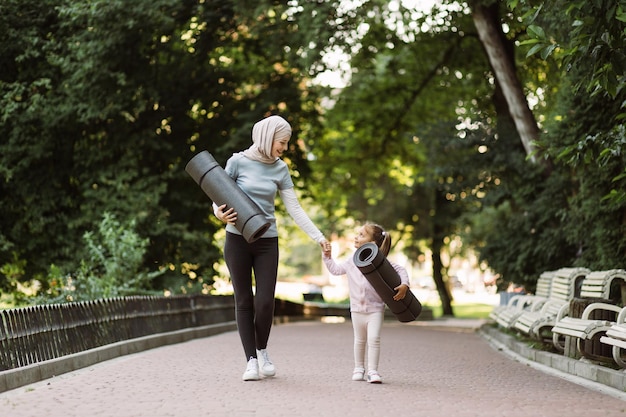 Mère musulmane marchant avec sa petite fille dans le parc tenant des tapis de yoga dans les mains