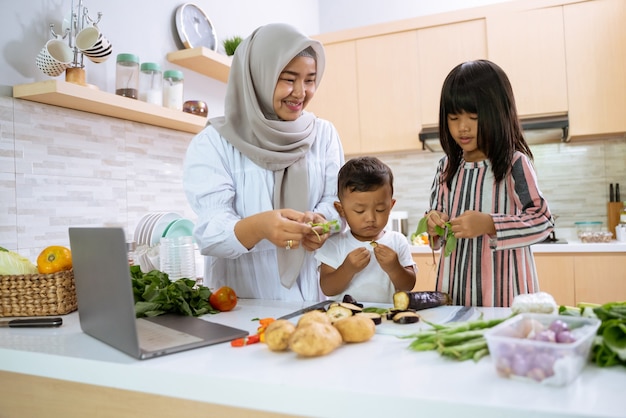 Mère musulmane regardant la vidéo de cuisine sur un ordinateur portable et préparer le dîner avec ses deux enfants dans la cuisine ensemble