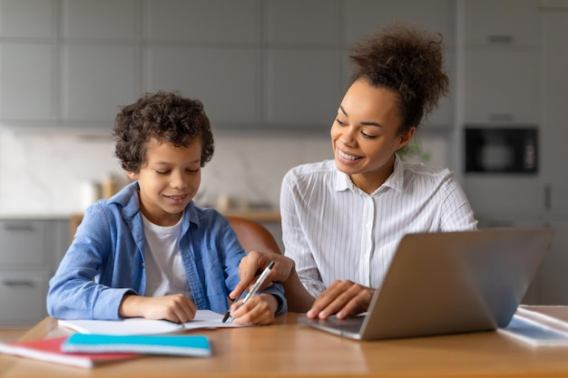 Une mère noire aide son fils à faire ses devoirs sur un ordinateur portable dans une cuisine bien éclairée