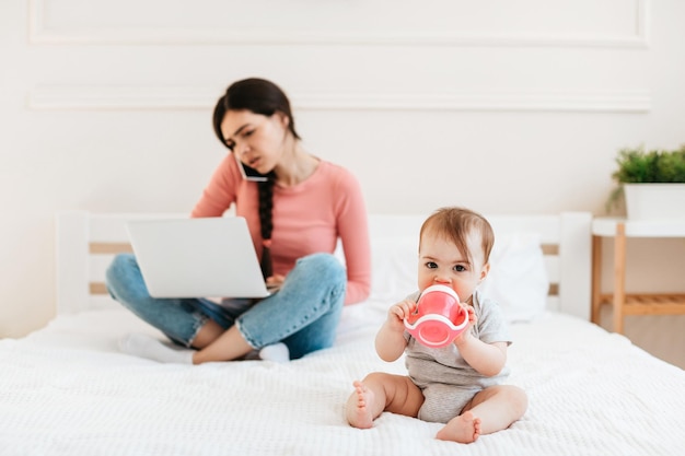 Mère occupée travaillant sur un ordinateur portable et parlant au téléphone pendant que son petit enfant boit de l'eau à la bouteille