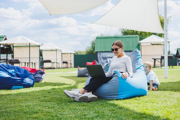 Photo mère avec ordinateur portable son fils jouant à l'extérieur sur la pelouse verte reposez-vous et travaillez avec un enfant dans le parc