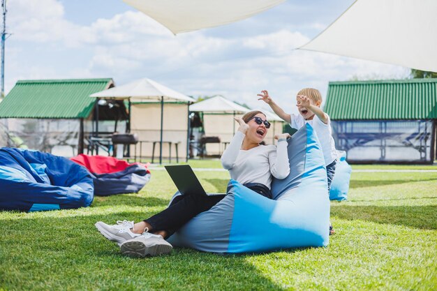 Photo mère avec ordinateur portable son fils jouant à l'extérieur sur la pelouse verte reposez-vous et travaillez avec un enfant dans le parc