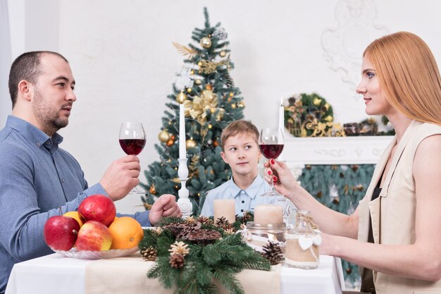 Mère, père et fils célèbrent Noël assis à une table, ramassant des verres de vin