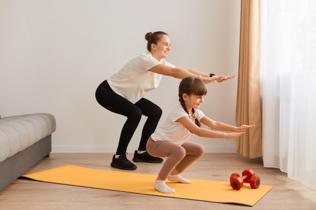 La mère et la petite fille pratiquant sont engagées dans la forme physique, le tapis d'exercice de sport à la maison, la femme avec l'enfant faisant des squats avec les mains tendues.