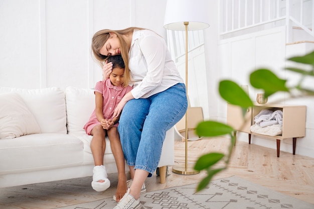 La mère a pitié de sa petite fille sur le canapé du salon. La mère et l'enfant de sexe féminin passent des loisirs ensemble dans leur maison, de bonnes relations, des soins parentaux et de l'amour