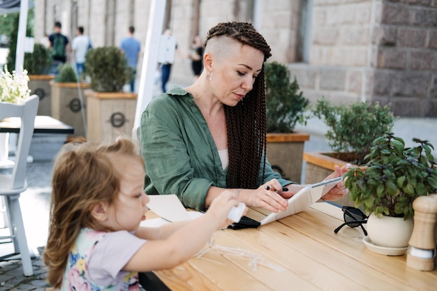 Mère et propriétaire d'entreprise prospère et occupée avec téléphone portable et tablette prenant son petit déjeuner avec elle