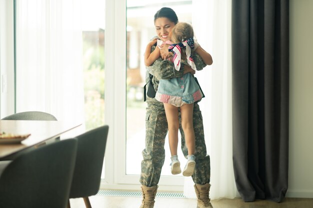 Mère de retour à la maison. Femme portant un uniforme militaire soulevant sa petite fille en rentrant à la maison