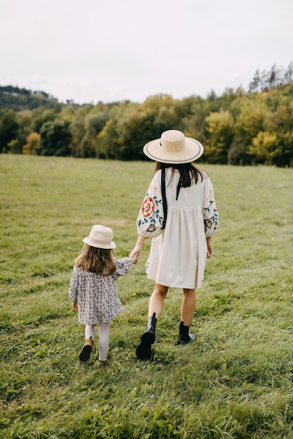 Une mère et sa petite fille marchent dans un champ ouvert, se tenant par la main, portant des chapeaux et des robes.