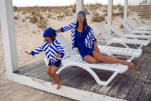 Mère et son bébé dans des vestes bleues à rayures marchent le long de la plage à côté d'un belvédère en bois