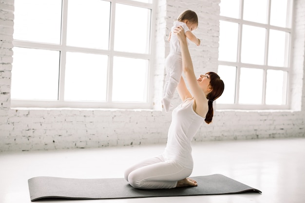 Une mère et son bébé font de la gymnastique, des exercices de yoga sur fond de mur blanc et de grandes fenêtres.