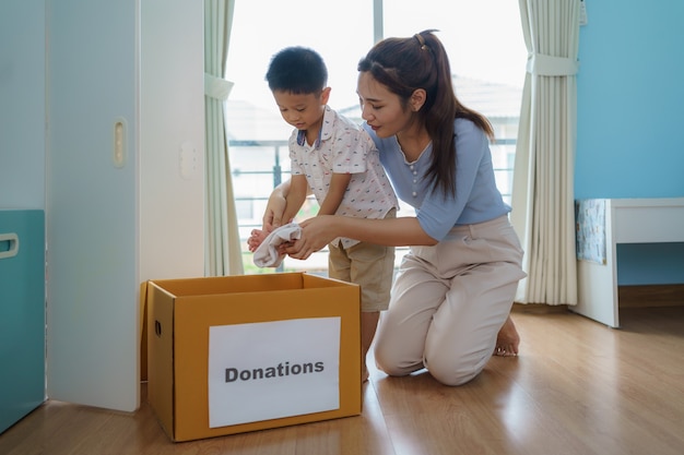 Une mère et son fils asiatiques se tiennent près d'un placard de vêtements dans le vestiaire, portant une boîte de vêtements donnée à apporter au centre de dons.