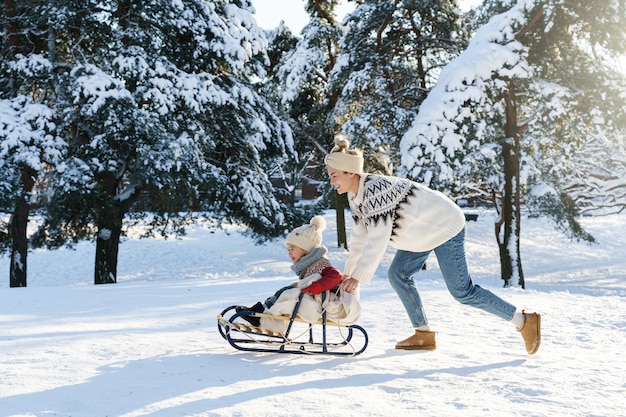 La mère et son mignon petit fils portant des chandails chauds s'amusent sur une colline de luge pendant la journée d'hiver ensoleillée