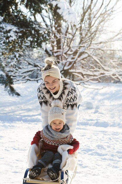 La mère et son mignon petit fils portant des chandails chauds s'amusent sur une colline de luge pendant la journée d'hiver ensoleillée