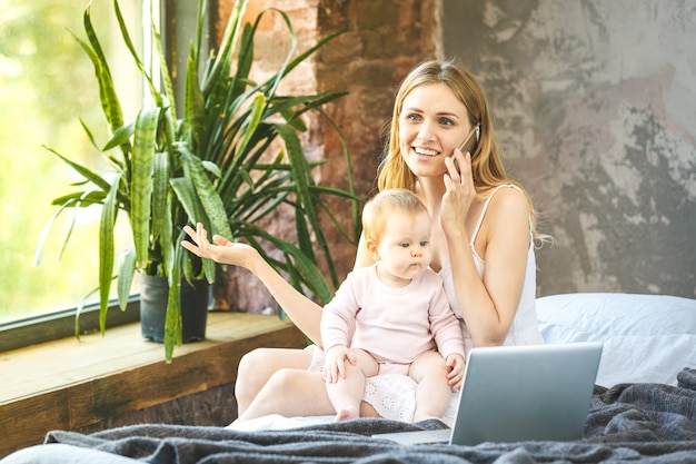 Mère et son petit bébé à la maison. Mère avec son bébé regardant quelque chose sur un ordinateur portable. Travail à domicile. Utilisation du téléphone.