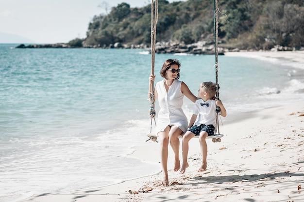 Mère avec son petit fils dans un t-shirt blanc et un nœud papillon s'amusant à se balancer sur une plage tropicale. Phuket. Thaïlande. Concept de vacances en famille