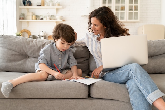 Une mère souriante à l'école à domicile aide son petit fils à écrire dans un ordinateur portable pendant le reste du travail à distance sur un ordinateur portable