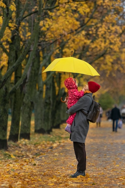 La mère tient un petit enfant dans ses bras alors qu'elle se tient sous un parapluie jaune dans un parc d'automne sur fond de feuillage jaune. Cadre vertical.