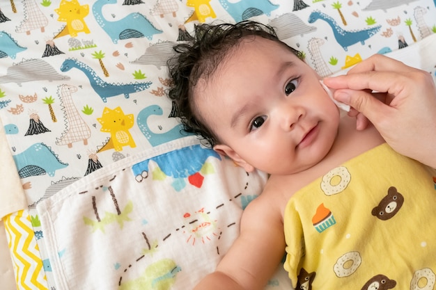 La mère utilise un coton-tige imbibé d'eau propre. Nettoyer le visage de bébé asiatique après le bain, petit bébé mignon regarde la caméra