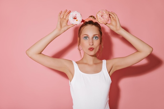 Merveilleuse dame aux cheveux blonds et maquillage moderne dans des vêtements légers regardant dans la caméra et tenant deux beignets sur fond isolé