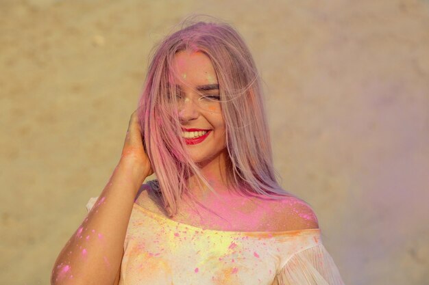 Merveilleuse femme blonde souriante jouant avec de la peinture sèche rose Holi dans le désert