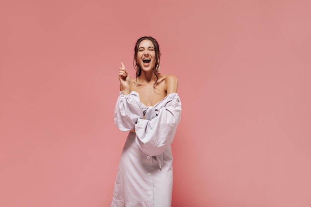 Merveilleuse fille bouclée en chemisier moderne rayé et jupe longue avec bouton blanc riant les yeux fermés sur mur rose