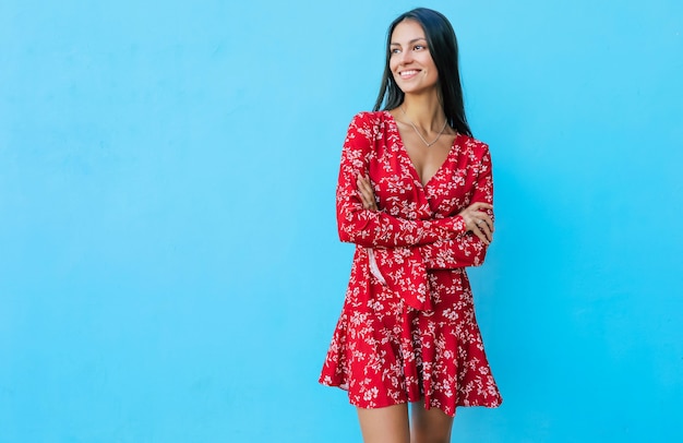 Merveilleuse fille en robe rouge pose sur fond bleu, tenant son poing droit près de son menton et avec sa main gauche pliée