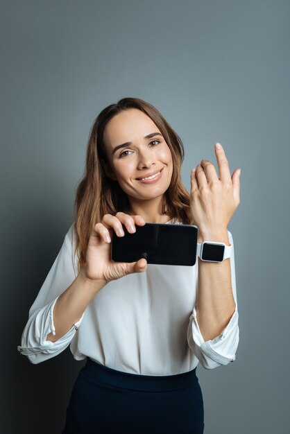 Mes appareils technologiques. Femme heureuse joyeuse positive tenant son smartphone et vous le montrant avec sa smartwatch en se tenant debout sur fond gris