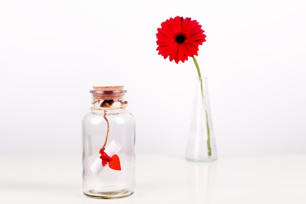 Message d'amour dans une bouteille rouleau de papier blanc avec fil rouge et fleur de gerbera rouge sur fond blanc