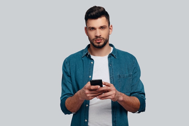 La messagerie texte. Charmant jeune homme utilisant son téléphone intelligent et regardant la caméra en se tenant debout sur fond gris