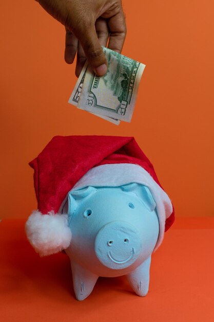 Mettre de l'argent de la main dans la tirelire avec le chapeau de père Noël de Noël isolé sur fond orange. Image conceptuelle.