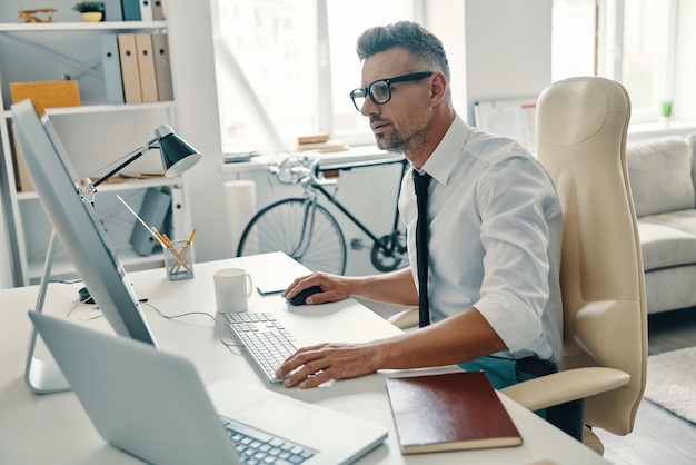 Mettre des idées dans quelque chose de réel. Gentil jeune homme en chemise et cravate travaillant à l'aide d'un ordinateur alors qu'il était assis au bureau