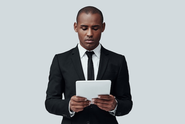 Mettre des idées dans quelque chose de réel. Homme africain réfléchi en tenue de soirée travaillant à l'aide d'une tablette numérique en se tenant debout sur fond gris