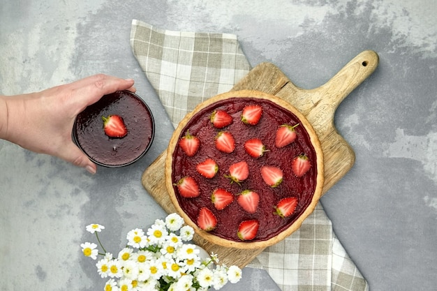 Mettre à la main de la confiture de fraises maison sur la table avec une tarte aux fraises et des fleurs de camomille à plat