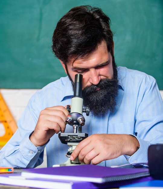 Mettre en place un microscope. L'enseignant s'assoit au bureau avec un microscope. Homme barbu hipster arrière-plan de tableau de classe occupé avec l'observation biologique. Professeur d'école à la recherche d'un microscope. Des recherches passionnantes.