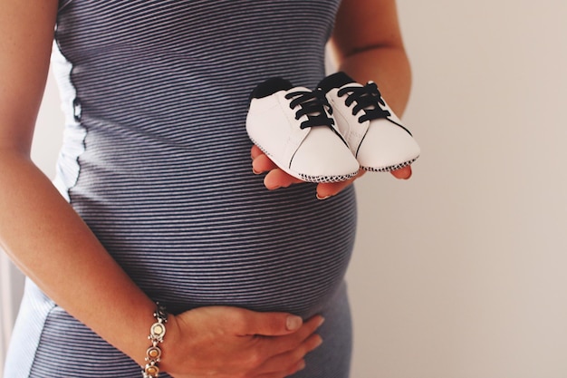 Midsection sur une femme enceinte tenant des chaussures alors qu'elle se tient contre le mur