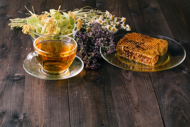Miel liquide avec nid d'abeille et tasse de thé à l'intérieur d'un tas d'herbes sèches sur une surface en bois blanche