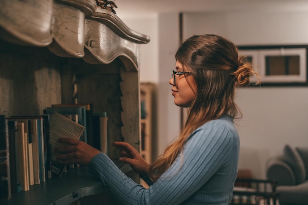 Un mignon et attrayant portant des lunettes lisant et regardant un livre intéressant à la maison à l'intérieur Jeune femme se détendre seule la nuit en lisant des livres