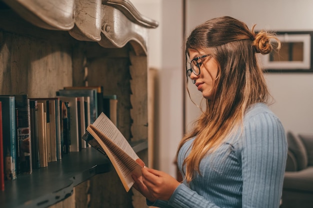 Un mignon et attrayant portant des lunettes lisant et regardant un livre intéressant à la maison à l'intérieur Jeune femme se détendre seule la nuit en lisant des livres