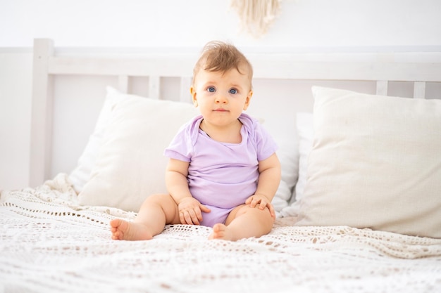 Un mignon bébé en bonne santé dans un body lilas rampe sur un lit avec des draps blancs dans la chambre de la maison un portrait d'un enfant un enfant regarde la caméra