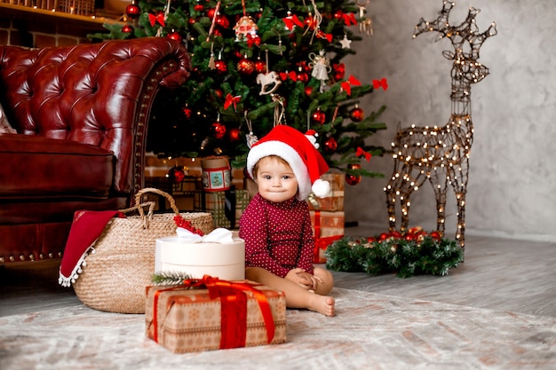 Mignon bébé Père Noël est assis à la maison près de l'arbre de Noël avec des cadeaux