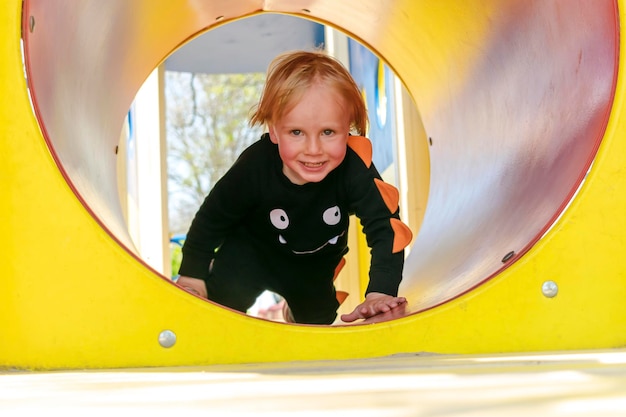 Mignon drôle blond petit jeune enfant en bas âge enfant garçon rampant grimper à travers un tunnel en plastique jaune dans l'aire de jeuxDéveloppement physique des enfants et concept de garderie de l'enfance