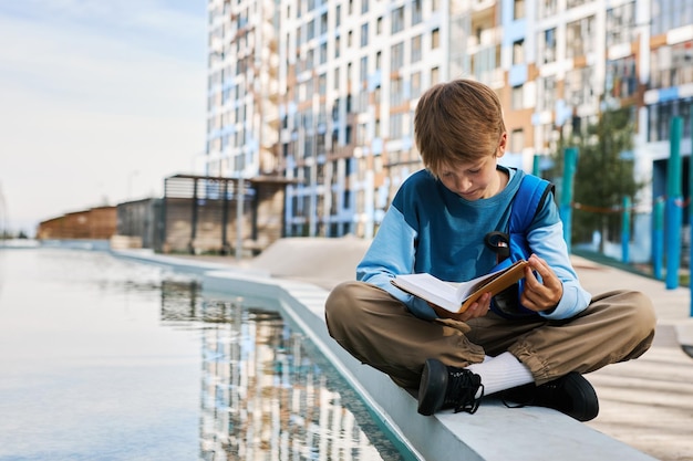 Mignon écolier diligent en tenue décontractée assis au bord de la rivière et lisant